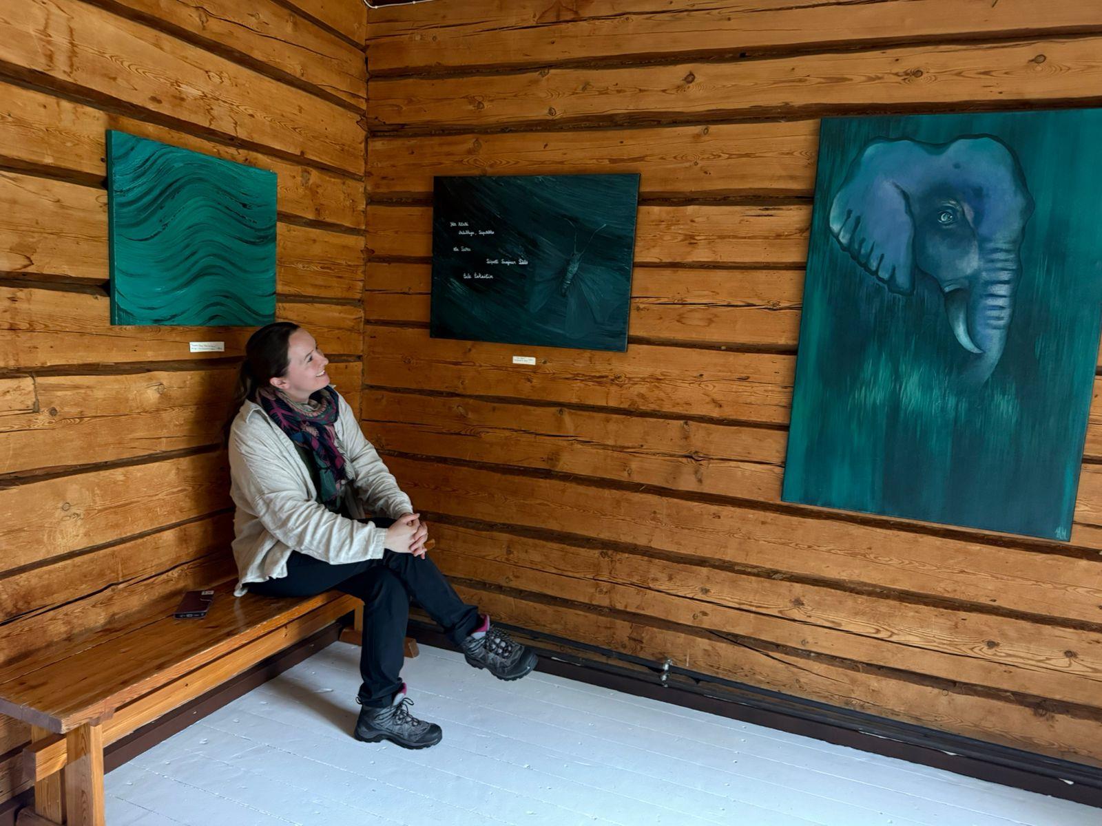 Visitor sitting on a bench in a wooden room with paintings at Lykynlampi