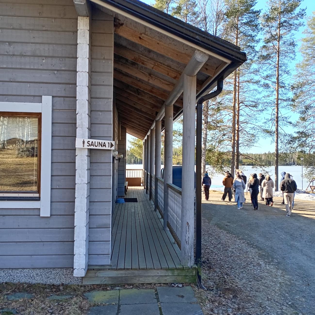 Wooden building with a sauna sign and people walking toward the lake at Koli Freetime